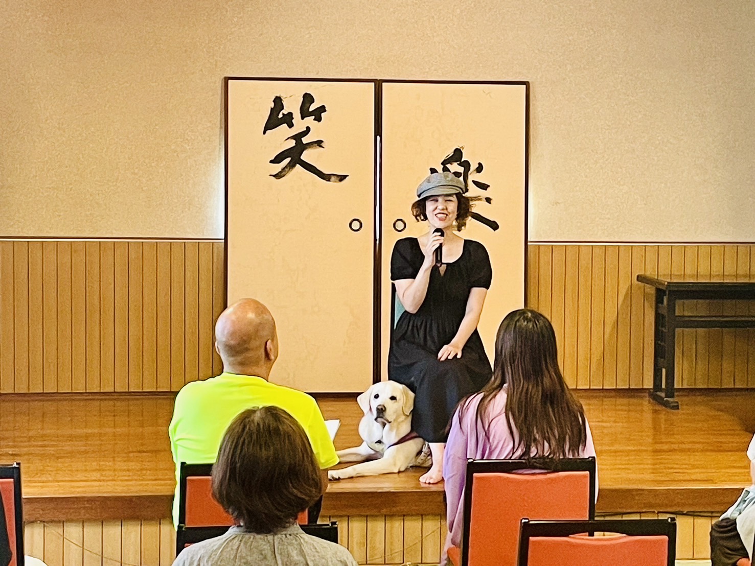 講演会のステージで盲導犬のヴィヴィッドくんと一緒に語っている浅田純子さんの写真（正面から撮影）